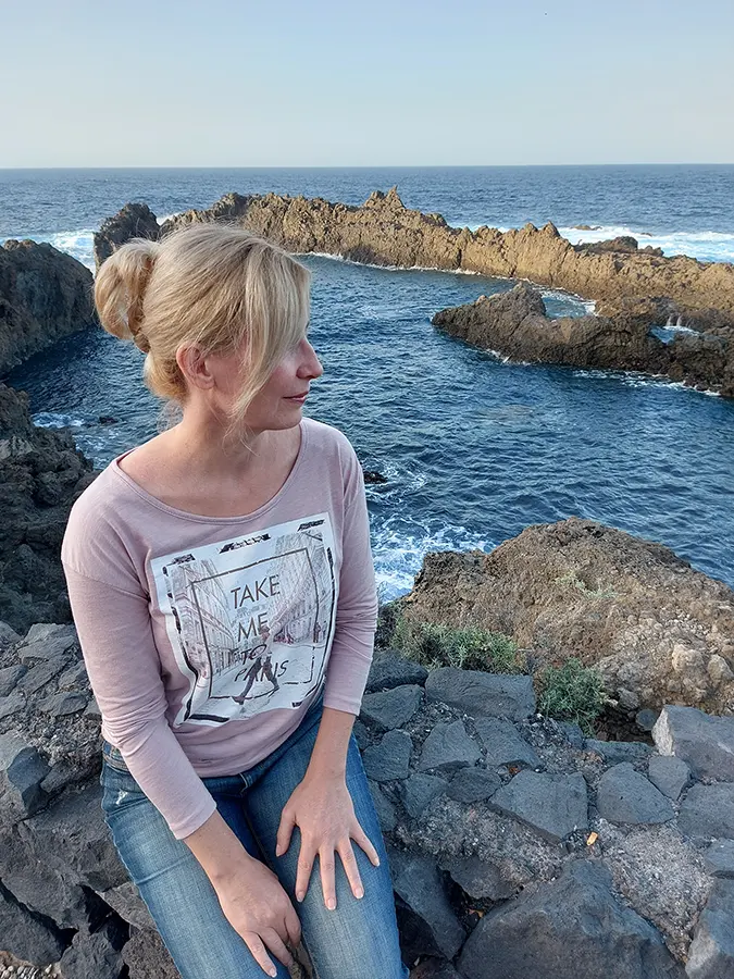 Góra Edina Tenerifén, a vízparton, köveken ül, nézi az óceánt és a sziklákat. Közben mosolyog. Szőke nő, haja feltűzve. Arca profilból látszik.